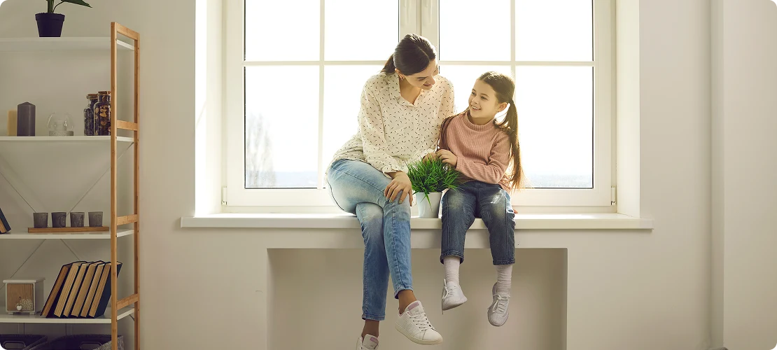 Мама с дочкой сидят на подоконнике у большого окна в светлой комнате